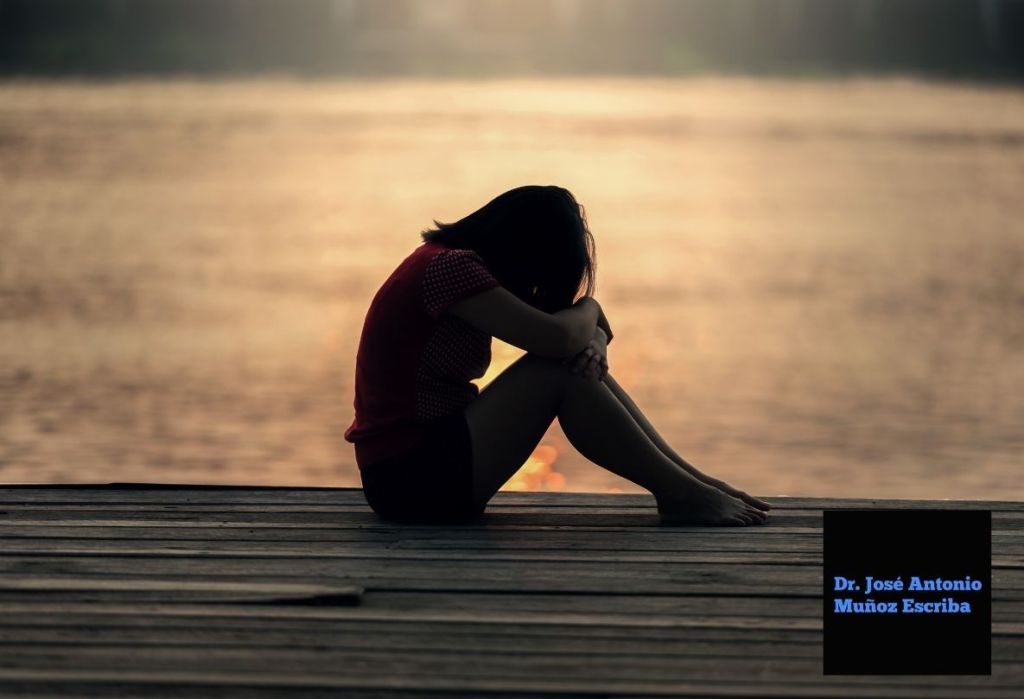 Mujer con la cabeza reposando en sus brazos, sentada en un muelle al atardecer. Esta imagen refleja el dolor mamario que presenta y utiliza esta posición para tratar de aliviarlo o hacerlo más tolerable